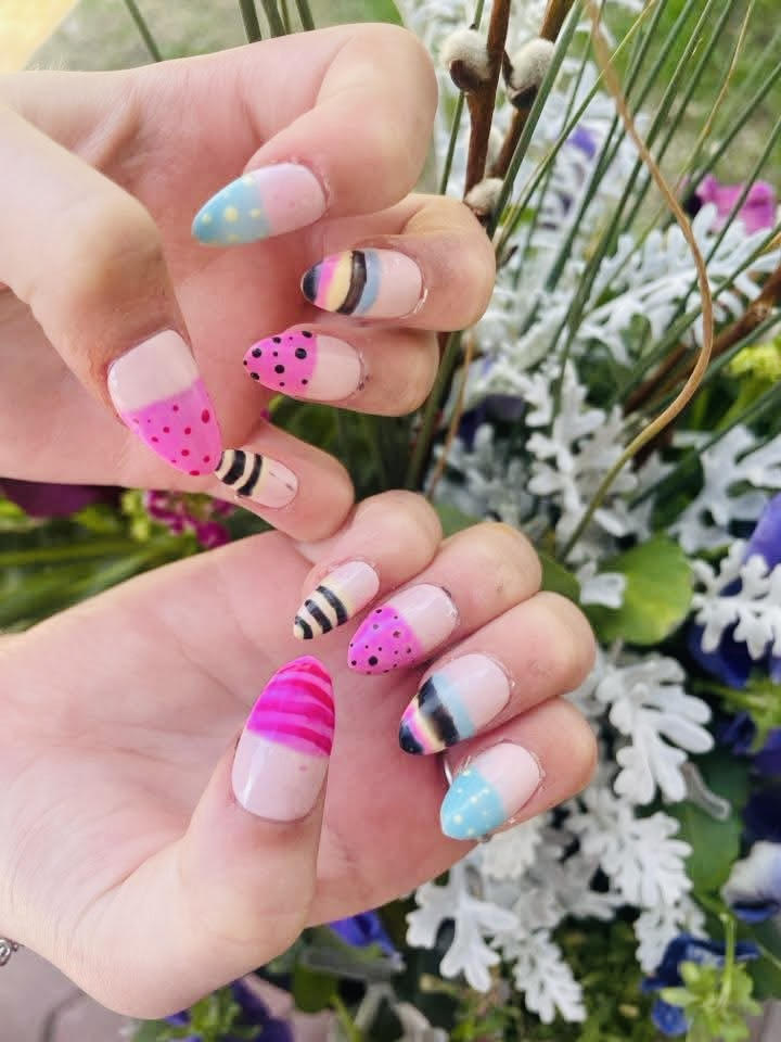 Close-up of hands showing colorful nail art with pastel tips, polka dots, stripes, and gradient designs against a garden background.